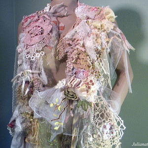 May include: A handmade, layered garment featuring a variety of textures and colors. The piece incorporates lace, ribbons, and floral accents in shades of pink, cream, and beige. Delicate tulle and crochet details add to the intricate design. The garment is displayed on a mannequin.