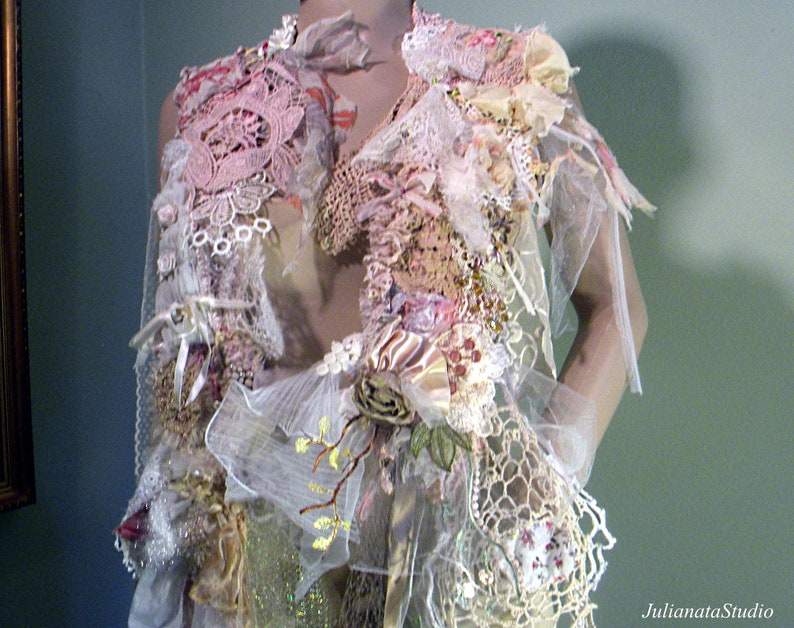 May include: A handmade, layered garment featuring a variety of textures and colors. The piece incorporates lace, ribbons, and floral accents in shades of pink, cream, and beige. Delicate tulle and crochet details add to the intricate design. The garment is displayed on a mannequin.