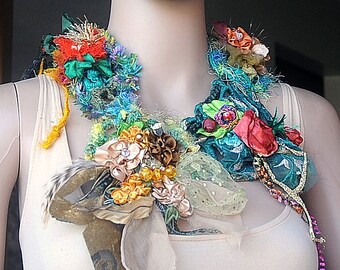 Puede incluir: Un collar llamativo hecho a mano y colorido. El collar presenta una variedad de texturas y colores, incluyendo turquesa, rojo, amarillo y dorado. Está adornado con flores de tela, cintas y cuentas. El collar se exhibe en un maniquí.