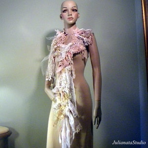 May include: A decorative scarf displayed on a mannequin. The scarf features layers of pink, white, and cream-colored fabrics, including lace and tulle. The design incorporates various textures and embellishments, creating a unique, handcrafted accessory.