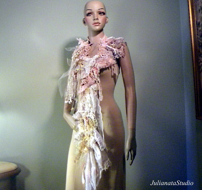 May include: A decorative scarf displayed on a mannequin. The scarf features layers of pink, white, and cream-colored fabrics, including lace and tulle. The design incorporates various textures and embellishments, creating a unique, handcrafted accessory.