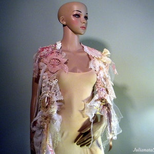 May include: A decorative, open-front garment with a floral and lace design. The piece features a mix of pink, cream, and gold tones, with intricate details and embellishments. The garment is displayed on a mannequin, showcasing its delicate and artistic craftsmanship.