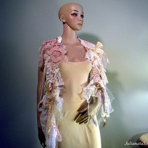 May include: A decorative, open-front garment with a layered design. The piece features a mix of lace, fabric, and floral embellishments in shades of pink, cream, and gold. The garment is displayed on a mannequin.