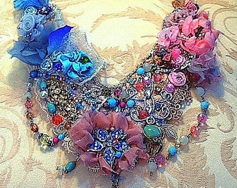 Puede incluir: Un collar de declaración de varias capas con una variedad de texturas y colores, incluyendo azul, rosa, turquesa y plata. El collar presenta flores, cuentas y encaje.