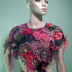 May include: A crocheted shawl featuring a floral design in shades of pink, purple, and green. The shawl is crafted with a variety of textures and materials, including crochet, beads, and fabric flowers.