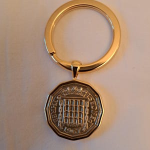 May include: Gold-toned keychain featuring a vintage Three Pence coin from 1961. The coin is octagonal with a grid design and text around the edge. The keychain ring and clasp are also gold-toned.