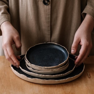 Puede incluir: Una pila de tres platos redondos de cerámica con una superficie interior negra mate y un borde de arcilla beige cruda y texturizada. Los platos presentan un diseño orgánico de bordes irregulares, mostrando una clara separación entre el centro oscuro y el borde claro y terroso de cada pieza individual.