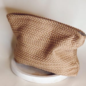 Könnte beinhalten: Eine beigefarbene gewebte Clutch mit Fischgrätmuster. Die Tasche hat einen Reißverschluss und liegt auf einer weißen, runden Oberfläche. Die Textur des Materials ist sichtbar. Die Tasche scheint leer zu sein.