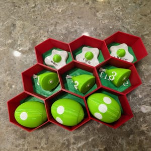 May include: A red and green educational toy with a honeycomb design. The toy contains green egg-shaped pieces with white dots, green cylinders, and white egg-shaped pieces with green centers. The words "one", "two", and "three" are visible.