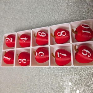 May include: A set of ten red apple-shaped number blocks, each with a white number from 1 to 10. The blocks are arranged in a white tray with individual compartments. Each apple has a small brown stem.