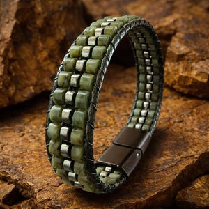May include: A green and silver bracelet with a magnetic clasp. The bracelet features rows of green stone beads and silver metal accents, set against a dark leather band. The clasp is a dark, metallic colour.
