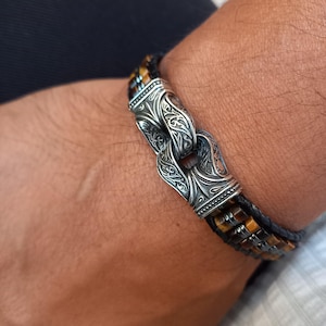 May include: A silver bracelet with an ornate clasp is shown. The bracelet has a braided leather band with brown and black details. The clasp is decorated with detailed patterns.