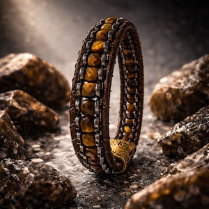May include: A brown leather bracelet featuring a central row of faceted golden-brown tiger eye beads flanked by dark metallic chains. The bracelet is secured with an ornate, cylindrical gold-toned metal clasp with intricate engraved detailing.