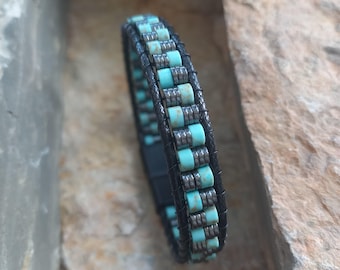 Op de afbeelding: Zwart leren armband met turquoise en zilverkleurige kralen. De armband heeft een multi-strand ontwerp met een magnetische sluiting. De turquoise kralen zijn cilindrisch en afgewisseld met zilverkleurige metalen kralen.