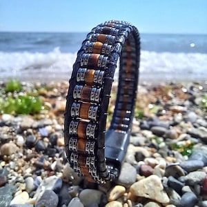 Puede incluir: Una pulsera de cuero negro con detalles marrones y plateados. La pulsera presenta un diseño trenzado con cuentas rectangulares marrones y elementos metálicos plateados. El fondo muestra una playa con guijarros y el mar.
