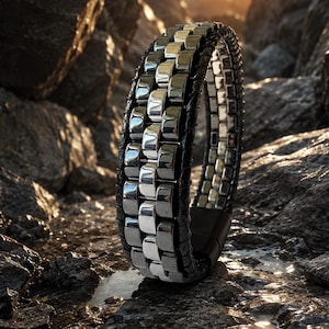 May include: A silver and black bracelet with a woven leather band. The bracelet features rectangular silver links and a magnetic clasp. The jewelry is displayed on a rocky surface.