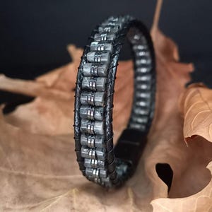 Pode incluir: Uma pulseira de couro preto com contas de pedra cinza e detalhes em metal prateado. A pulseira é feita de várias fileiras de contas retangulares, conectadas por arame fino, e tem um fecho magnético. A pulseira é exibida em uma folha marrom.