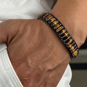 Puede incluir: Primer plano de una pulsera con piedras rectangulares de ojo de tigre. Las piedras están engarzadas en una banda de cuero negro con detalles metálicos plateados. La pulsera se lleva en la muñeca.