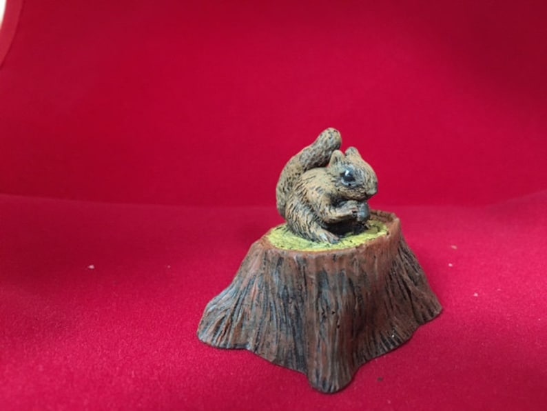 May include: A small, brown squirrel figurine sitting on a tree stump. The squirrel is holding a nut in its paws and has a bushy tail.