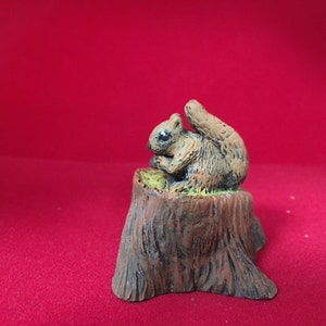 May include: A small, brown squirrel figurine sitting on a tree stump. The squirrel is holding a nut in its paws and has a bushy tail.