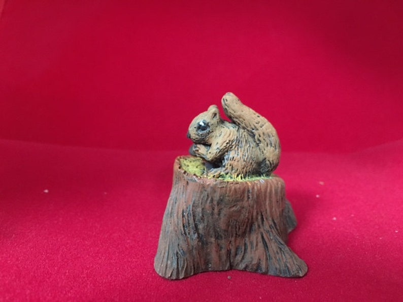 May include: A small, brown squirrel figurine sitting on a tree stump. The squirrel is holding a nut in its paws and has a bushy tail.