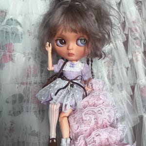 May include: A custom Blythe doll with grey hair in a bun and a lavender dress. The doll has large eyes and a small brown purse. It is wearing white lace socks and brown shoes. A pink lace dress is next to the doll. The doll is approximately 20 cm tall.