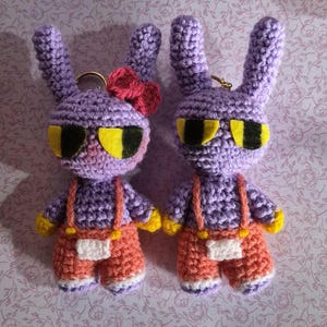 May include: Two crocheted purple rabbit keychains with long ears, yellow felt eyes, and orange overalls. One rabbit features a small pink bow on its ear. Both figures have yellow hands and white accents on their feet and pockets, attached to gold rings.