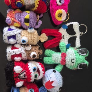 May include: A collection of ten small, handmade crochet keychains featuring various colourful characters with yarn textures, felt details, and metal rings. The figures include diverse shapes, expressions, and vibrant colour combinations like red, blue, green, purple, and white.