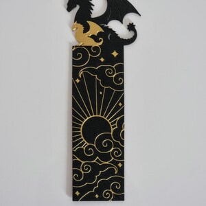 May include: Black and gold bookmark featuring a dragon silhouette with a sun and clouds design. The dragon is perched on a cloud with its wings spread out. The sun is in the center of the bookmark with rays emanating from it. The clouds are stylized and have a swirling pattern.