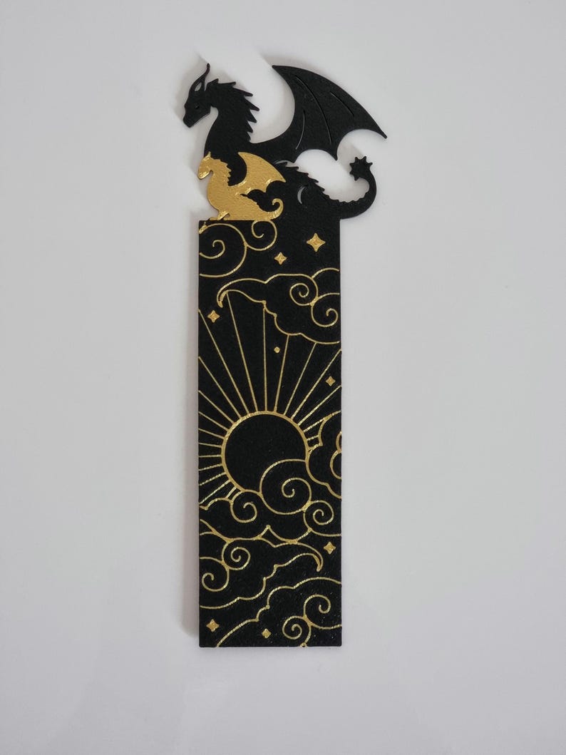 May include: Black and gold bookmark featuring a dragon silhouette with a sun and clouds design. The dragon is perched on a cloud with its wings spread out. The sun is in the center of the bookmark with rays emanating from it. The clouds are stylized and have a swirling pattern.