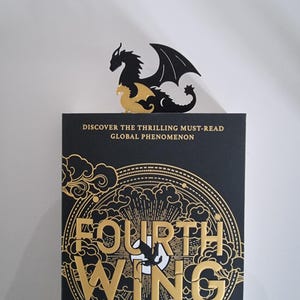 May include: A black and gold book cover with the title "Fourth Wing" in gold lettering. The cover features a stylized dragon design with clouds and a circle.