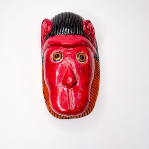 May include: A red wooden mask with black hair, orange eyes, and a carved nose and mouth.
