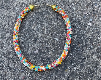 May include: A colorful beaded necklace with gold accents. The necklace is made of small, round beads in various colors, including red, orange, yellow, green, blue, and purple. The beads are strung together in a random pattern, creating a unique and eye-catching design.