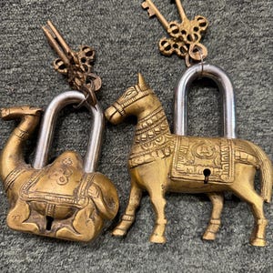 Puede incluir: Dos candados de latón con arcos plateados. Un candado tiene forma de camello y el otro de caballo, ambos con detalles grabados intrincados y bocallaves. Cada candado incluye una llave de latón ornamentada a juego unida al arco del candado.