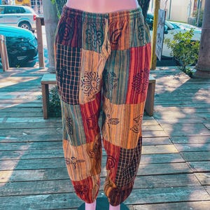 Könnte beinhalten: Patchwork-Hose mit elastischem Bund und Kordelzug. Das Kleidungsstück besteht aus verschiedenen rechteckigen Stoffbahnen in Orange-, Rot-, Braun- und Grüntönen, die mit schwarzen floralen und geometrischen Mustern bedruckt sind.