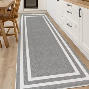 May include: A rectangular gray rug with a white border in a kitchen setting. The rug has a textured appearance and is placed on a light-colored wooden floor. The kitchen features white cabinets and a wooden table.