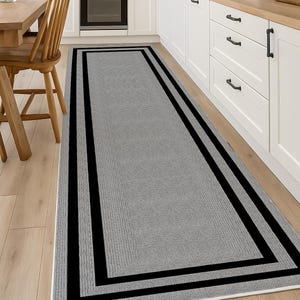 Peut inclure: Un long tapis de couloir rectangulaire doté d'un panneau central gris clair texturé, encadré par une bordure noire audacieuse et un fin liseré blanc. Le tapis présente une structure tissée à profil bas posée sur un sol en bois clair.