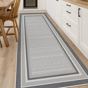 Può includere: Un tappeto passatoia rettangolare caratterizzato da un centro grigio chiaro con un sottile motivo testurizzato, incorniciato da bordi multipli in varie tonalità di grigio scuro e bianco, creando un design geometrico a strati.