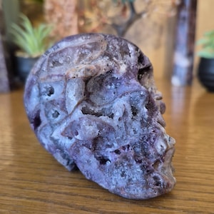 May include: A purple and white crystal skull carved from amethyst. The skull is detailed with intricate carvings and has a rough, textured surface.