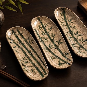 Puede incluir: Tres platos de cerámica ovalados y alargados con un esmalte color crema moteado y tallos y hojas de bambú de color verde oscuro pintados a mano. Cada plato tiene un acabado liso y brillante con un borde ligeramente elevado para servir.