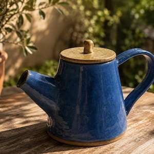 Puede incluir: Una tetera de cerámica con un cuerpo de esmalte azul profundo brillante y un asa curva. La tapa es de un color beige moteado que contrasta con un pequeño pomo central. La base de la tetera no está esmaltada, dejando ver el material de arcilla.