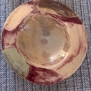 Op de afbeelding: Een ronde keramische schaal met een glanzende afwerking en abstracte, handgeschilderde penseelstreken in tinten diep bordeaux, gedempt bruin en zacht beige. Het midden van de schaal is licht verdiept, wat een ondiepe kom met een glad oppervlak vormt.