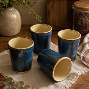 May include: A set of four ceramic tumblers featuring a deep blue exterior glaze with a smooth, cream-colored interior. Three of the tumblers stand upright, while one is positioned on its side, showcasing the cylindrical shape and the contrast between the dark blue outer finish and the light-colored inner surface.