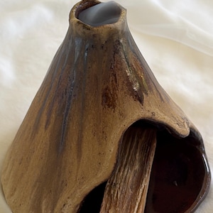 May include: A cone-shaped ceramic incense burner with a textured tan and brown exterior glaze. The burner features a hollow interior chamber with a dark brown glazed base and a single stick of light brown wood resting inside the opening.
