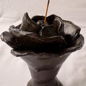 May include: A dark brown, glazed ceramic incense holder shaped like a blooming rose. The sculpture features a flared, pedestal-style base and layered, textured petals. A single, thin, brown incense stick is inserted into the center of the flower.