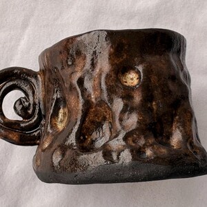 Puede incluir: Una taza de cerámica de color marrón oscuro con una superficie brillante, irregular y texturizada. La taza presenta un asa gruesa y distintiva en forma de espiral. La base de la taza es plana y oscura, contrastando con el acabado de esmalte marrón.