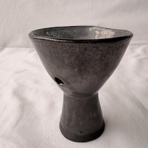 May include: A dark gray ceramic chalice with a wide, flared rim and a narrow, cylindrical base. The surface features a mottled, metallic finish with a small, circular opening located on the side of the bowl section, near the stem.