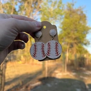 May include: Baseball-themed earrings. These dangle earrings feature a white baseball design with red stitching. The earrings are round and have a stud top. The background is blurred, with trees and sunlight.