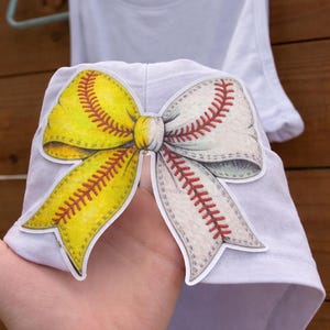 May include: A white tank top with a softball and baseball bow design. The bow features a yellow softball side and a white baseball side, both with red stitching details. The tank top is hanging on a hanger.