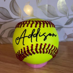 May include: A softball with the name "Addison" in black script. The softball is yellow with red rhinestones along the seams. It sits on a pink base.
